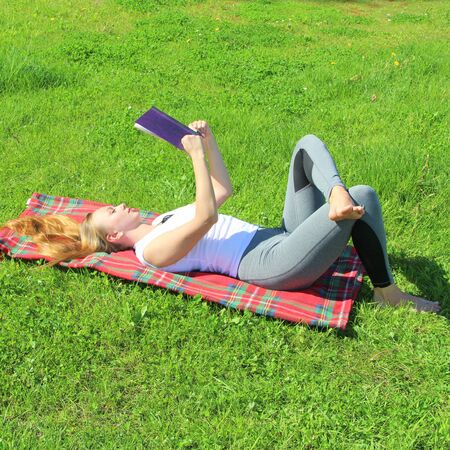 A beautiful young white girl in a white T-shirt and with long hair lies on a red plaid, on green grass, on the lawn and read book.の写真素材
