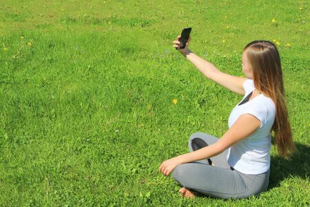 A beautiful young white girl in a white t-shirt and with long hair sitting on green grass, on the lawn and takes a selfie.の写真素材