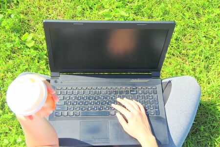 The girl sits on the green grass on the lawn, holding a black laptop on her lap. Laptop close-up.の写真素材