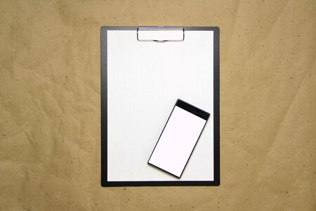 The tablet with a clip for the clip of paper with a white sheet a4 lies against the background of craft brown crumpled paper. Above is a mobile phone with a white screen. Place for text and layout for design.の写真素材