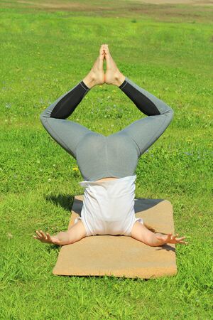 Young woman of European appearance does yoga in summer nature. Woman standing in pose closed angle headstand, badhakona shirshasana. High quality photo for web and print with empty space for text and design.の写真素材