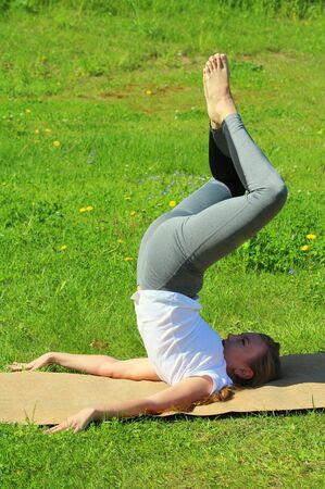 Young woman of European appearance does yoga in summer nature. Woman standing in pose closed angle headstand, badhakona shirshasana. High quality photo for web and print with empty space for text and design.の写真素材