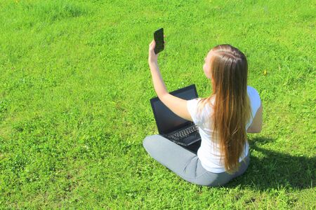 A beautiful young white girl in a white t-shirt and with long hair sitting on green grass, on the lawn and working behind a black laptop with a mobile phone and takes a selfie.の写真素材