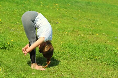 Young woman of European appearance does yoga in summer nature. Woman standing in forward tilt pose with hand lock, baddha hasta uttanasana. High quality photo for web and print with empty space designの写真素材