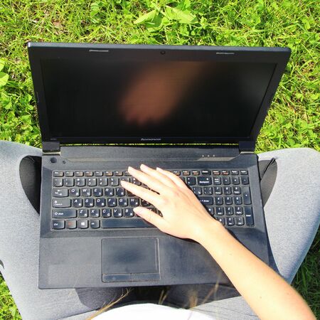 The girl sits on the green grass on the lawn, holding a black laptop on her lap. Laptop close-up.の写真素材