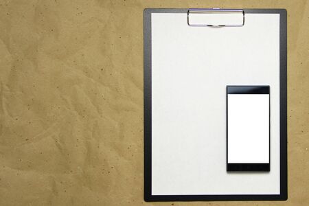 The tablet with a clip for the clip of paper with a white sheet a4 lies against the background of craft brown crumpled paper. Above is a mobile phone with a white screen. Place for text and layout for design.の写真素材