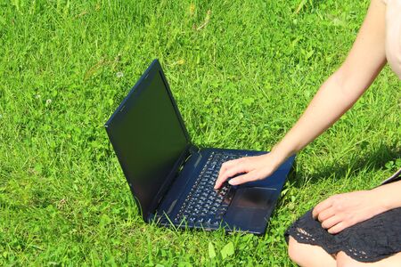 A beautiful young white girl in a pink jacket and black skirt and with long hair sitting on green grass, on the lawn and working behind a black laptop.の写真素材