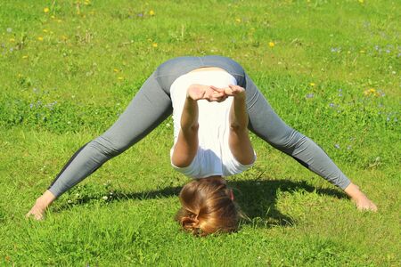 Young woman of European appearance does yoga in summer nature. Woman stands head to foot pose, shishra angushtha yogasana. High quality photo for web and print with empty space for text and design.の写真素材