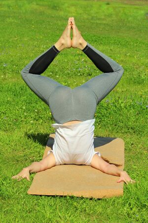 Young woman of European appearance does yoga in summer nature. Woman standing in pose closed angle headstand, badhakona shirshasana. High quality photo for web and print with empty space for text and design.の写真素材