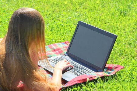 A beautiful young white girl in a white T-shirt and with long hair lies on a red plaid, on green grass, on the lawn and working behind a black laptop.の写真素材