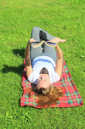 A beautiful young white girl in a white T-shirt and with long hair lies on a red plaid, on green grass, on the lawn and read book.の写真素材