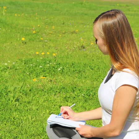 A beautiful young white girl in a white t-shirt and with long hair sitting on green grass, on the lawn and writes with a pen in a notebook.の写真素材