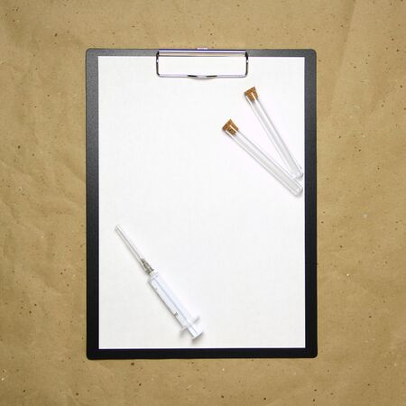 The tablet with a clip for the clip of paper with a white sheet a4 lies against the background of craft brown crumpled paper. On top is a medical syringe and two transparent empty tubes with stoppers. Place for text and layout for design.の写真素材