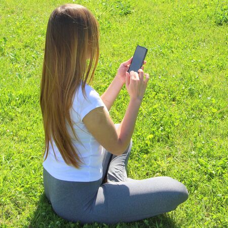 A young beautiful girl of European appearance in a white T-shirt with long blond hair sits on green grass, on the lawn with a mobile phone in her hands, flips through social networks, works in the phone.の写真素材