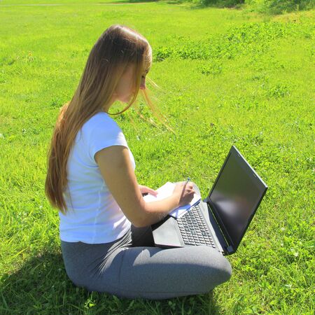 A beautiful young white girl in a white t-shirt and with long hair sitting on green grass, on the lawn and working behind a black laptop and writes with a pen in a notebook.の写真素材