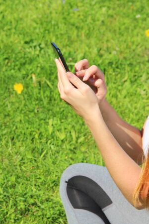 A young beautiful girl of European appearance in a white T-shirt with long blond hair sits on green grass, on the lawn with a mobile phone in her hands, flips through social networks, works in the phone.の写真素材
