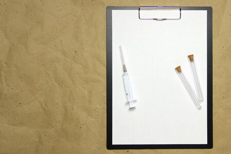 The tablet with a clip for the clip of paper with a white sheet a4 lies against the background of craft brown crumpled paper. On top is a medical syringe and two transparent empty tubes with stoppers. Place for text and layout for design.の写真素材