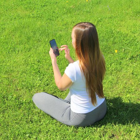 A young beautiful girl of European appearance in a white T-shirt with long blond hair sits on green grass, on the lawn with a mobile phone in her hands, flips through social networks, works in the phone.の写真素材