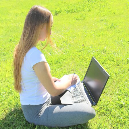 A beautiful young white girl in a white t-shirt and with long hair sitting on green grass, on the lawn and working behind a black laptop and writes with a pen in a notebook.の写真素材