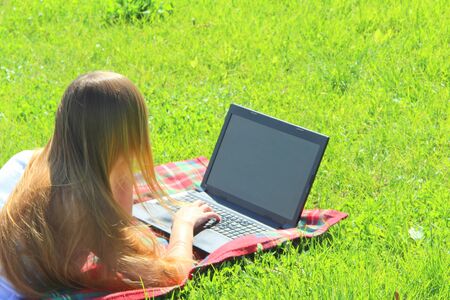 A beautiful young white girl in a white T-shirt and with long hair lies on a red plaid, on green grass, on the lawn and working behind a black laptop.の写真素材