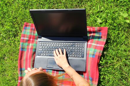 A beautiful young white girl in a white T-shirt and with long hair lies on a red plaid, on green grass, on the lawn and working behind a black laptop.の写真素材