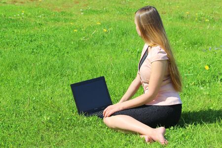 A beautiful young white girl in a pink jacket and black skirt and with long hair sitting on green grass, on the lawn and working behind a black laptop.の写真素材
