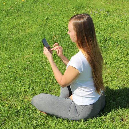 A young beautiful girl of European appearance in a white T-shirt with long blond hair sits on green grass, on the lawn with a mobile phone in her hands, flips through social networks, works in the phone.の写真素材