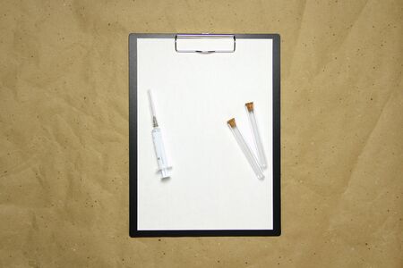 The tablet with a clip for the clip of paper with a white sheet a4 lies against the background of craft brown crumpled paper. On top is a medical syringe and two transparent empty tubes with stoppers. Place for text and layout for design.の写真素材