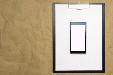 The tablet with a clip for the clip of paper with a white sheet a4 lies against the background of craft brown crumpled paper. Above is a mobile phone with a white screen. Place for text and layout for design.の写真素材