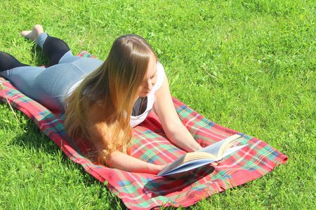 A beautiful young white girl in a white T-shirt and with long hair lies on a red plaid, on green grass, on the lawn and read book.の写真素材