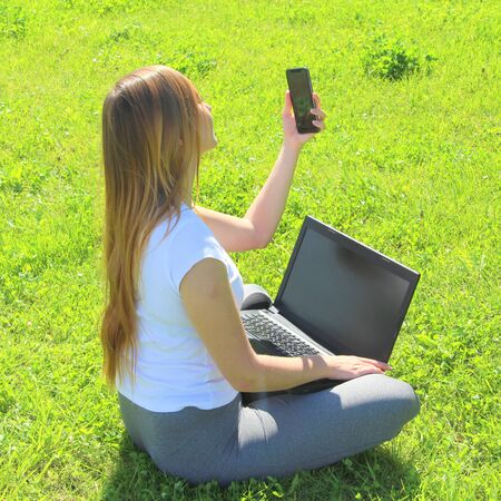 A beautiful young white girl in a white t-shirt and with long hair sitting on green grass, on the lawn and working behind a black laptop with a mobile phone and takes a selfie.の写真素材