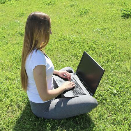 A beautiful young white girl in a white t-shirt and with long hair sitting on green grass, on the lawn and working behind a black laptop.の写真素材