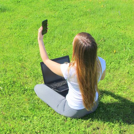 A beautiful young white girl in a white t-shirt and with long hair sitting on green grass, on the lawn and working behind a black laptop with a mobile phone and takes a selfie.の写真素材