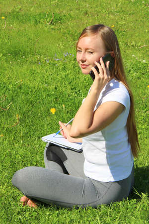 A beautiful young white girl in a white t-shirt and with long hair sitting on green grass, on the lawn and working behind a black laptop and talking on a cell phone and writes with a pen in a notebook.の写真素材
