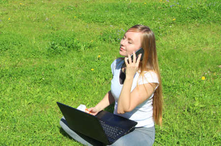 A beautiful young white girl in a white t-shirt and with long hair sitting on green grass, on the lawn and working behind a black laptop and talking on a cell phone.の写真素材