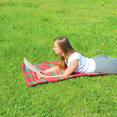 A beautiful young white girl in a white T-shirt and with long hair lies on a red plaid, on green grass, on the lawn and read book.の写真素材