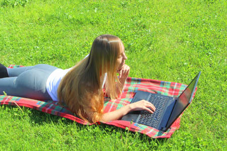 A beautiful young white girl in a white T-shirt and with long hair lies on a red plaid, on green grass, on the lawn and working behind a black laptop.の写真素材