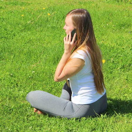 A beautiful young white girl in a white t-shirt and with long hair sitting on green grass, on the lawn and working behind a black laptop and talking on a cell phone and writes with a pen in a notebook.の写真素材