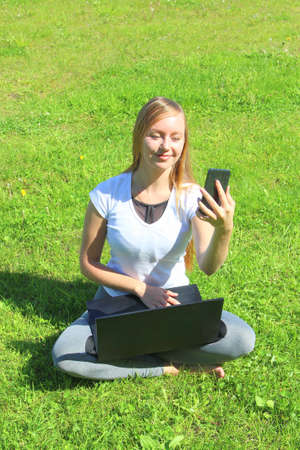 A beautiful young white girl in a white t-shirt and with long hair sitting on green grass, on the lawn and working behind a black laptop with a mobile phone and takes a selfie.の写真素材