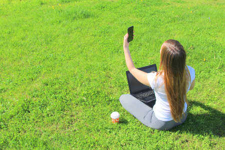 A beautiful young white girl in a white t-shirt and with long hair sitting on green grass, on the lawn and working behind a black laptop with a mobile phone and takes a selfie.の写真素材