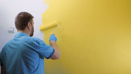 Man paints white wall with roller in his right hand in yellow. He is wearing plaid blue shirt and...