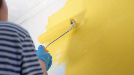 Girl paints wall with roller in her right hand in yellow. She is wearing striped shirt and blue glovesの写真素材