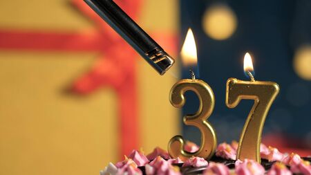 Birthday cake number 37 golden candles burning by lighter, background gift yellow box tied up with...