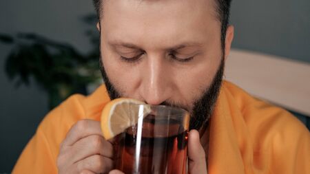 Front view guy is drinking black tea. Attractive young man is ill and drinks tea with lemon while sitting in bed. Cold, flu, sore throat, runny nose, acute respiratory disease concept. Close-upの写真素材