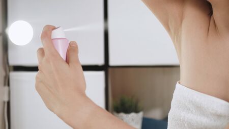 Girl uses deodorant. Young attractive woman in white towel in bathroom with her left hand sprays...