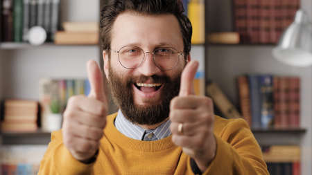 Man thumb up. Positive laughing bearded man in glasses in office or apartment room looking at camera and shows his two hands thumb up. Close-upの写真素材