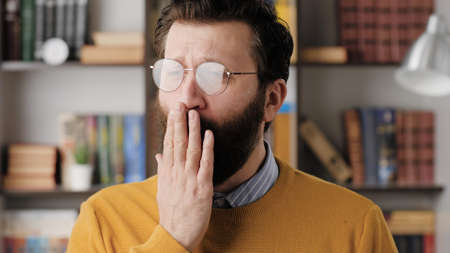 Man yawns. Tired sad bearded man in glasses in office or apartment room yawns, he covers his mouth...