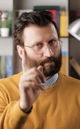 Man threatens with his finger. Serious frowning bearded man in glasses in office or apartment room looking at camera and points menacingly with his index finger. Close upの写真素材