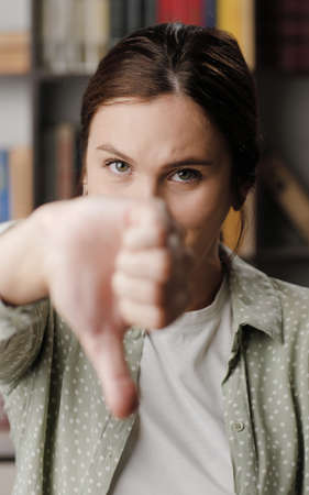 Woman thumb down. Frowning serious woman in office or apartment room looking at camera raises her hand and shows her thumb down. Medium shotの写真素材