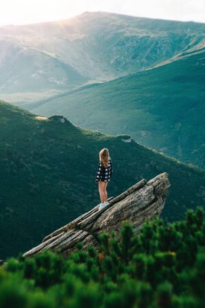 Beautiful young blonde girl standing on the rock in front of big mountain.